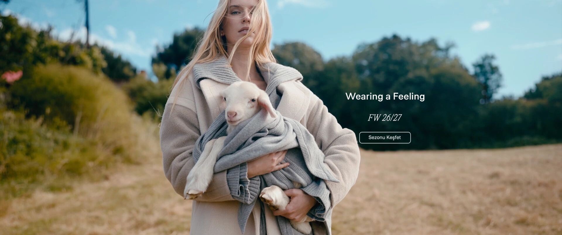Wearing a Feeling, FW 26/27 — kollarında kuzu taşıyan, bej kaban ve gri triko giymiş kadın
