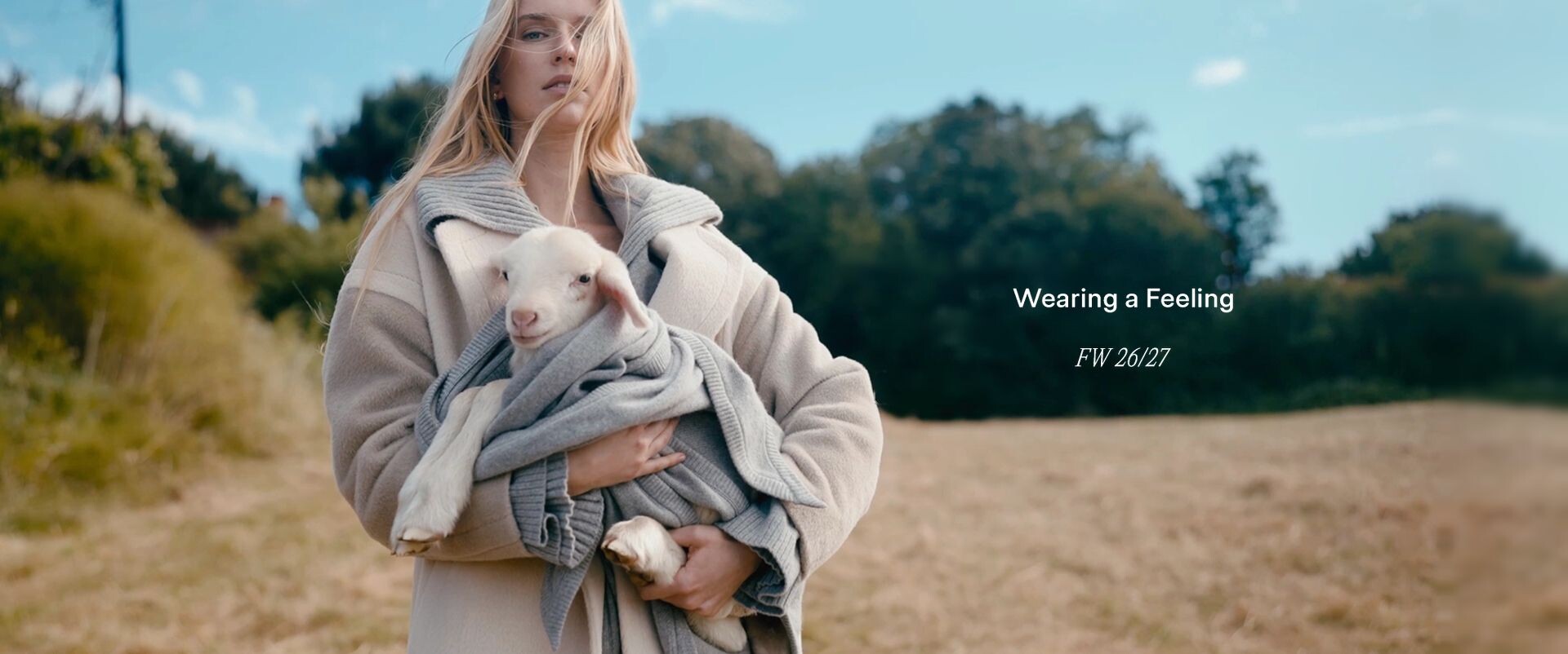 Wearing a Feeling, FW 26/27 — kollarında kuzu taşıyan, bej kaban ve gri triko giymiş kadın